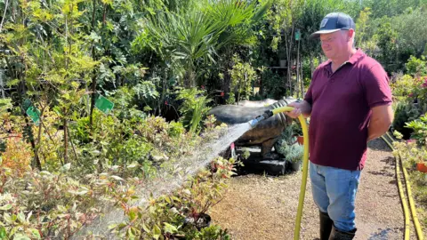 John Devine/BBC John Cutteridge is holding a yellow hosepipe to spray some flowers outside in a garden centre. He is wearing a blue baseball cap and a mauve t-shirt with blue jeans and wellington boots.