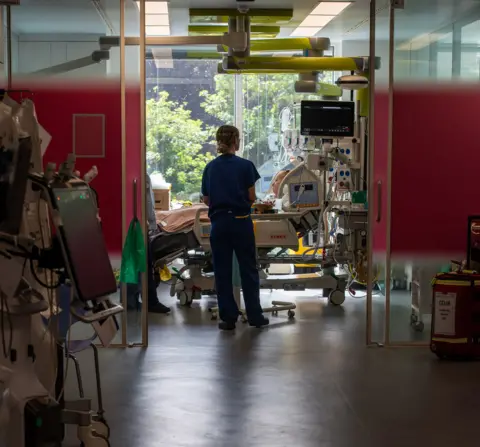 Hollie, 29, is propped up on pillows in her hospital bed in an ICU room inside King's College Hospital, wearing a hospital gown with tubes attached to her nose, and her mother sits by her bedside (unseen) while a nurse checks her.