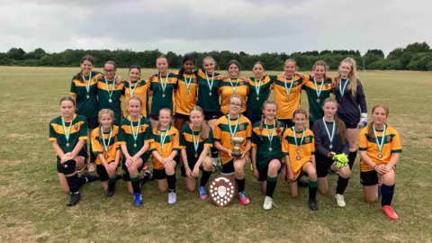 A team of young girls pictured with their trophies on a football pitch. The girls are arrange in two rows with one row standing and the other kneeling. The kits are yellow and green.