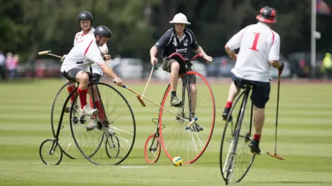 Penny Farthing Club Four players ride penny farthing bicycles across a grass pitch, striking a yellow ball with wooden mallets