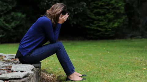 PA Media A woman dressed in blue with her head in her hands, while sat outside in a garden