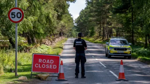 PA Media Closed road. A police officer is standing on a road with his back to the camera. He is next to a red and white Road Closed sign and two orange and white traffic cones. There is a police car further down the road