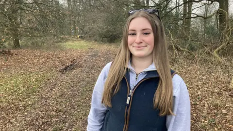 Jazz White smiling at the camera while standing in woodland. She is wearing a grey top and blue gilet. She has long light brown hair and has sunglasses on the top of her head. 