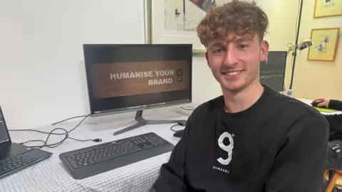 A man sits at a computer desk and smiles into the camera. A screen with the words 'Humanise Your Brand' can be seen behind him alongside a keyboard and laptop.