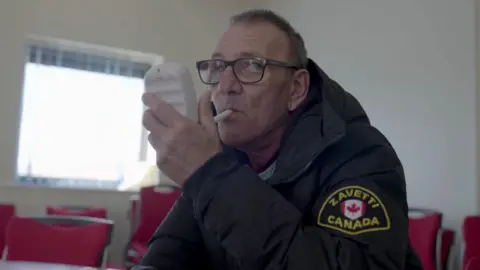 A screenshot from the 'Quitters' which shows Andy, a man who looks to be in his 60s, wearing glasses with a black frame. He is holding a small white machine in his hand, with a tube in his mouth as he blows in to it. He is white, has short grey hair and is wearing a black puffer coat with a Canada badge on the left sleeve. He is sitting in a room with red chairs and a large square window in the background. 
