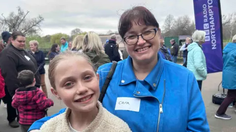 Kate Bradbrook/BBC Mother and daughter, Cara and Freya. On the left is Freya, an 11-year-old girl with blonde hair which is scraped back. She is looking directly at the camera and smiling while wearing a cream coloured jacket. Next to her is her mum Cara who also has her hair scraped back and is wearing a blue zip up suede jacket with a white name tag which says Cara on it. She is also looking directly at the camera and smiling. Behind them are crowds of people and a purple sign with white writing which says BBC Radio Northampton, on it.