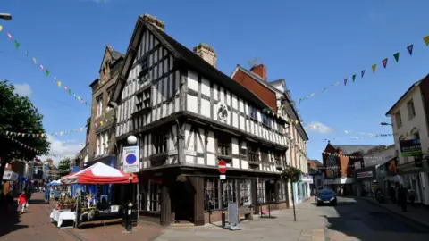 The corner of a two-storey black and white timbered building with pedestrianised streets running down both sides and market stalls with red and white awnings on the left hand side. There is bunting hanging over both streets.