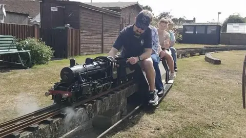 Romney Marsh Model Engineering Society People sat with one of the train models at the Romney Marsh Model Engineering Society. Four people can be seen in the image, with the man at the front making adjustments to the model.