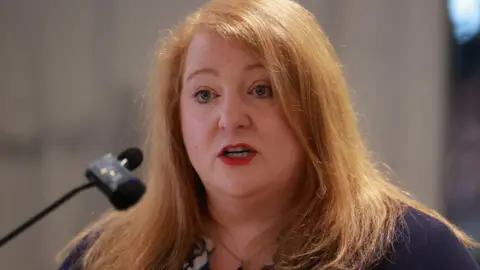 Justice Minister Naomi Long speaks down a microphone. She has red hair and red lipstick and is wearing a navy top.