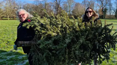 Sue Ryder Two volunteers - one on the left wearing glasses and a black jacket with a red strip- holding a Christmas tree with another lady standing on the other side- holding the top of the tree 