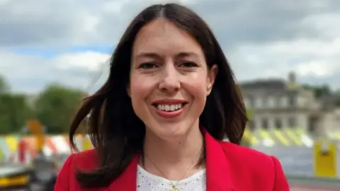 Paul Moseley/BBC Alice Macdonald is standing outside at the top of Norwich Market, which can be seen in the background behind her. She has brunette hair and is smiling while looking at the camera. She is wearing a white top, gold necklace and red blazer.