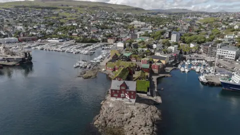 Drone footage of Tórshavn marina, with government buildings, some of which have grass roofs, visible in the foreground. A number of boats can also be seen in the picture