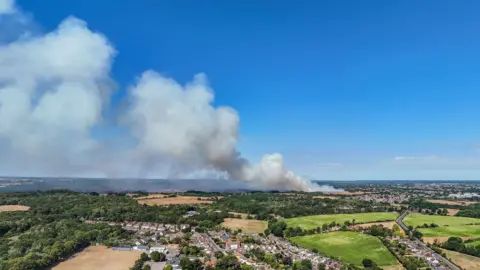Liam Winters A bird's eye drone view of fields, trees and houses. In the background white smoke billows into the sky. The sky is blue.