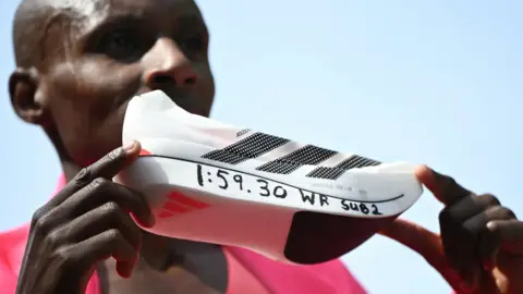 A white Adidas running shoe is held up so that its side is central to the image. It has the three black stripes on it for the Adidas logo and the text 1:59:30 WR sub 2 is written along the base of the sole. The two hands of marathon world record holder Sabastian Sawe can be seen grasping the shoe lightly between fingers, with Sawe's blurred face, covered by the shoe, is partially seen in the background