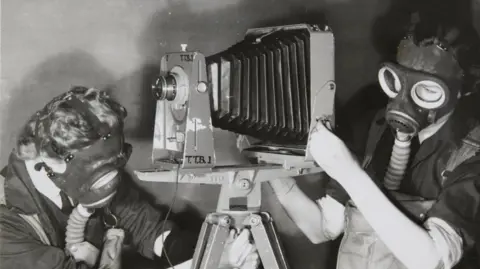 Historic England Archive Two female trainees practising photography whilst wearing gas masks to simulate working under active service conditions. There are wearing unfiformed jackets and overalls. The photo is black and white.