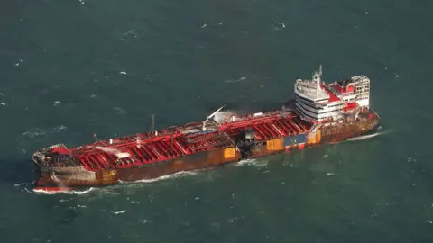 Vista aérea de Media PA de la Stena Immaculada con cubiertas rojas y superestructura blanca con un agujero en el casco y las marcas de quemaduras alrededor del costado de la nave