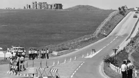 PA Media A 1985 black and white photo showing police with cones and road closed signs near Stonehenge before the Battle of the Beanfield