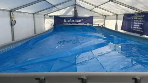A large blue pool inside a white tent with dark blue signs on the side, one of them reading "Embrace"