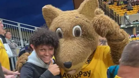 Family handout Harleigh is smiling with his thumb stuck up and arm round a football mascot at a Wolves match. He has long, curly black hair. The Wolves mascot is a big furry wolf wearing a gold top.