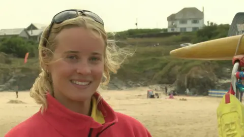 Erin Archibald smiling at the camera. She is wearing a red fleecy jacket and is standing on a beach. There is a house on top of the hill on the right. To her side is a yellow lifeboard.