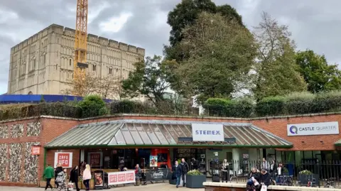 Exterior view of one of the entrances to the Castle Quarter shopping centre, with a cafe outside, and Norwich Castle visible on top of the shopping centre.