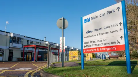 Exterior of the two-storey QE hospital front. A white sign to the right foreground indicates where to go for the main car park, breast care unit, Macmillan Care and Treatment Centre, West wing, main entrance and A&E. Two ambulances are parked outside the hospital entrance, which has a red-framed porch.