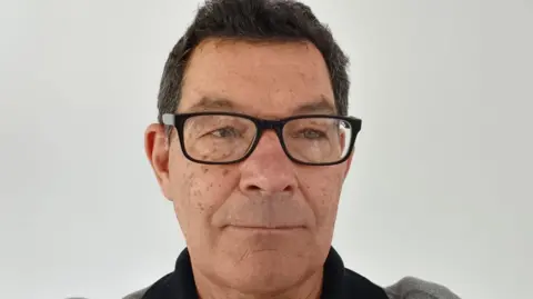 Robin Hughes A man with dark hair and black-framed glasses stands as he poses for the camera against a light background