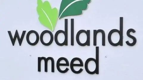 A sign reads Woodlands Meed in black font against a white background. 