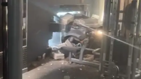 The front of a silver or grey car can be seen coming through a hole in the wall of the gym. Bricks and debris are lying around. Some gym equipment can be seen, including one apparatus which is right by where the car ended up.