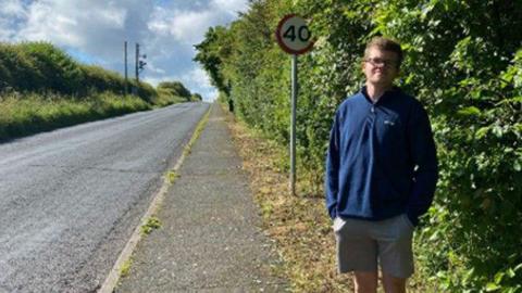Adam Myers next to the tidied up hedge with a 40mph speed limit sign and the road in the background