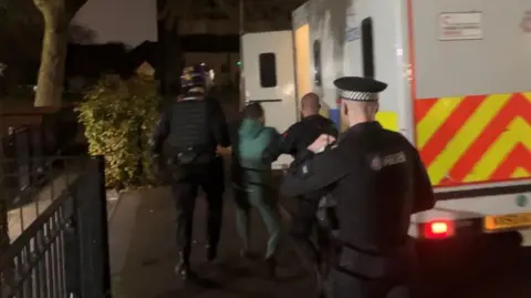 Photograph of a man being taken into custody with officers leading him into a police van.