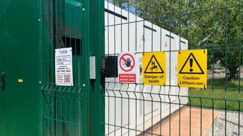 Wire fencing around the battery energy storage system with a white, red and black sign saying authorised access only and two yellow and black signs - one saying danger of death and the other saying caution lithium-ion.