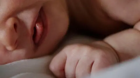 Generic close-up image of part of a newborn baby's face and hand. The baby lies on its side on a white blanket.