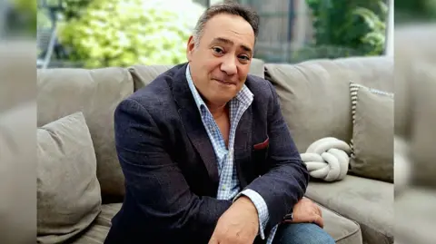 YMCA A picture of Joseph Main sat on a brown sofa with some brown cushions. There is also a cream knot cushion near the corner of the sofa. He is wearing a navy blue suit jacket with a blue chequered shirt that is slightly buttoned down. He is also wearing blue jeans. 