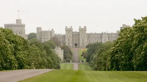 Getty Images A long, tree-lined path leads to the grand entrance of Windsor Castle, with multiple towers and battlements. There's greenery on either side of the path and the sky above is overcast.