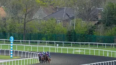 Horses and riders rounding a bend on a racetrack at Kempton Park, with green fencing, trees and houses visible beyond the course