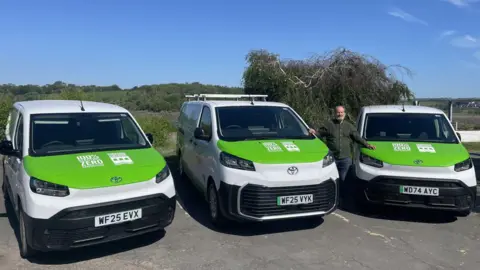 Councillor Peter Hames standing with three electric vans that are white with green bonnets.