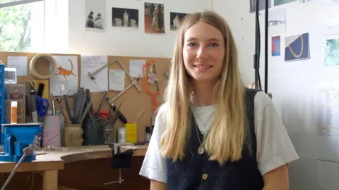 The image shows Iona Hall wearing a white t-shirt underneath a denim pinafore, with silver chains layered around her neck. She has long blonde hair and is smiling at the camera, sitting at a desk full of tools used to make jewellery.