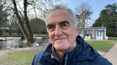 Martin Heath/BBC Ash Smith with short white hair looking at the camera and wearing a blue anorak. There is a river behind him, with trees on the bank and a cafe in the background.