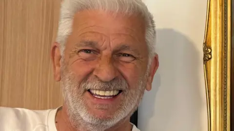 Stewart Woods, with white hair and a short white beard, smiles for the camera. He's wearing a white T shirt and is standing in front of a wall with the edge of a gold picture or mirror frame visible.