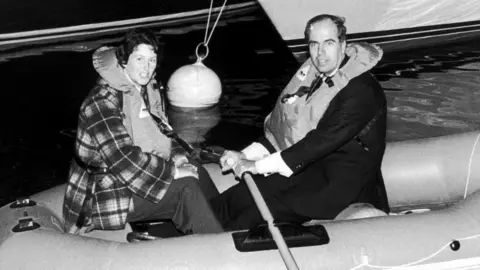 Getty Images Black and white photo of Maralyn and Maurice Bailey in a rubber dinghy and wearing life vests to relive their ordeal at the London Boat Show in 1974