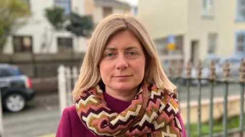 BBC Lyndsay Feltham is a woman with blonde hair. She is looking at the camera. She is wearing a pink, cream and yellow scarf over a pink coat. She is standing in front of a street with iron railings.