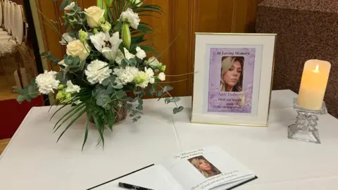 A bunch of flowers and a photograph of a woman are among the items sitting on a table. A book and pen and lit candle can also be seen. All items are atop a white cloth.