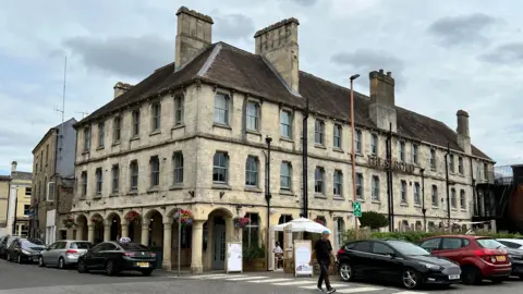 A large building made of lightly-coloured stone, with cars parked around it and a sign reading "The Stroud" on the right hand side.