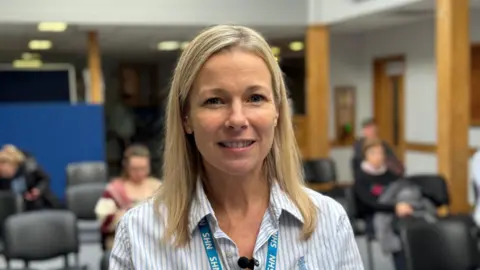 Josh McLaughlin/BBC Steph Williams stand in the health centre waiting room. She has shoulder length blond hair and is wearing a white shirt with blue stripes and a blue NHS lanyard.