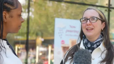 Two women face each other during an outdoor interview, with one holding a microphone and blurred background of a sign and trees.