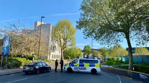 Police tape closes off the road. Two officers can be seen stood next to a police vehicle. The area is leafy with a pale building in the background. 