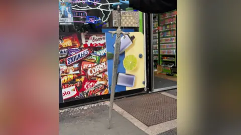 A large medieval-looking sword is propped up by an officer wearing blue gloves outside a shop.