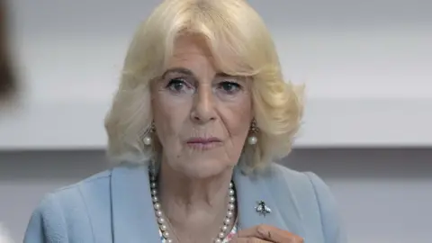 PA Media Queen Camilla wearing a blue dress and a pearl necklace and earrings looking towards the camera. 