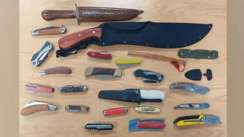 Twenty-four knives of various sizes resting on a table. Some are large, some are small and many are pocket knives. 